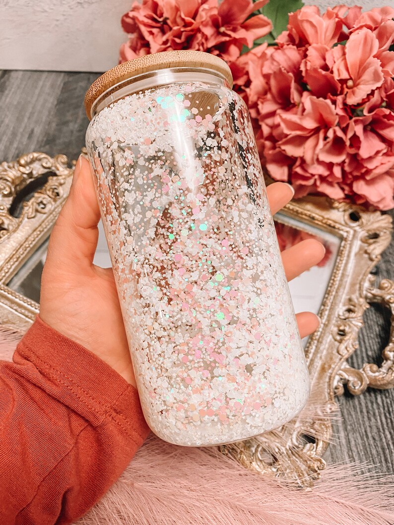 Peut inclure: Une bouteille en verre transparente avec un couvercle en bambou remplie de paillettes blanches et roses iris&eacute;es.