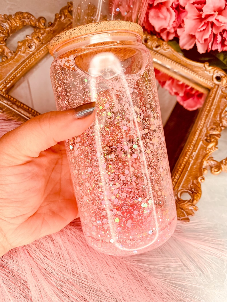 Peut inclure: Un bocal en verre transparent contenant des paillettes roses et un liquide. Le bocal a un couvercle en bois et est tenu par une main.