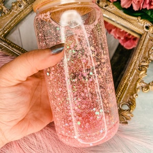 Peut inclure: Un bocal en verre transparent avec un couvercle en bambou rempli de paillettes roses. Le bocal est tenu dans une main.
