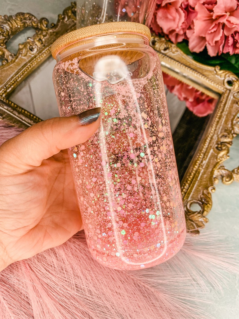 Peut inclure: Un bocal en verre transparent avec un couvercle en bambou rempli de paillettes roses. Le bocal est tenu dans une main.