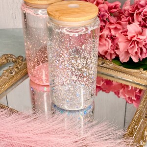 Peut inclure: Deux bocaux en verre transparent avec des couvercles en bambou remplis de paillettes iris&eacute;es. Les bocaux sont pos&eacute;s sur un plateau dor&eacute; avec un boa en plumes rose au premier plan.