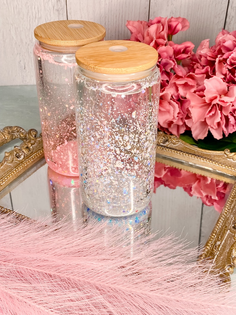 Peut inclure: Deux bocaux en verre transparent avec des couvercles en bambou remplis de paillettes iris&eacute;es. Les bocaux sont pos&eacute;s sur un plateau dor&eacute; avec un boa en plumes rose au premier plan.
