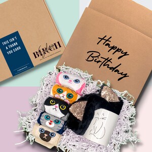 May include: A brown cardboard box with the text "Happy Birthday" on the front. The box is open and filled with white shredded paper. Inside the box are five pairs of cat-themed socks, a cat-shaped mug, and a black and gold cat ear headband.