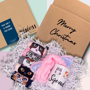 May include: A gift box filled with white shredded paper, containing a pink cat ear headband, a white mug with the word "Squad" and cat illustrations, and a set of five pairs of cat-themed socks. The box is brown with the text "Merry Christmas" printed on it.
