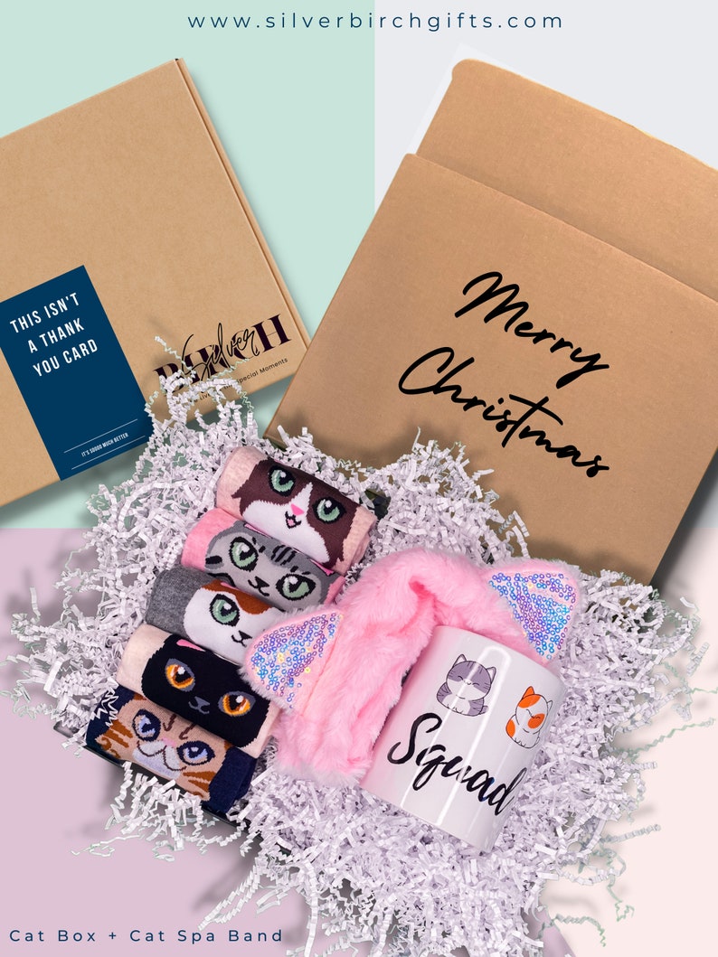May include: A gift box filled with white shredded paper, containing a pink cat ear headband, a white mug with the word "Squad" and cat illustrations, and a set of five pairs of cat-themed socks. The box is brown with the text "Merry Christmas" printed on it.