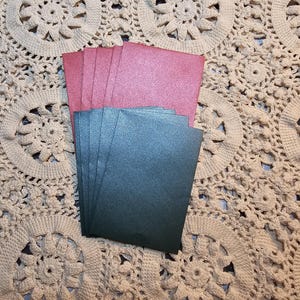 May include: A stack of pink and a stack of dark teal rectangular cards. The cards have a glittery, textured surface. The cards are arranged on a cream-colored, crocheted doily with a circular pattern.