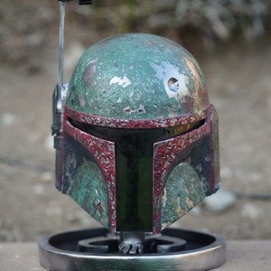 Puede incluir: Un casco de Boba Fett verde y rojo desgastado con una antena negra. El casco está en un soporte plateado.