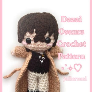 Op de afbeelding: Handgemaakte gehaakte pop van een personage met bruin haar, een beige jas en een zwarte outfit. De pop heeft grote zwarte ogen en is voorzien van het label "Dazai Osamu Crochet Pattern" met het julierumi-logo.