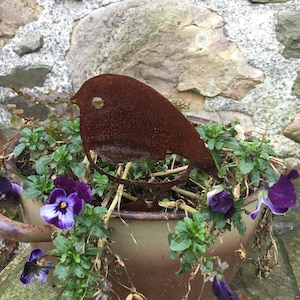 Könnte beinhalten: Eine rostige Vogelskulptur aus Metall sitzt in einem braunen Keramiktopf, der mit violetten Stiefmütterchen und grünem Laub gefüllt ist. Der Topf steht auf einer Steinoberfläche.