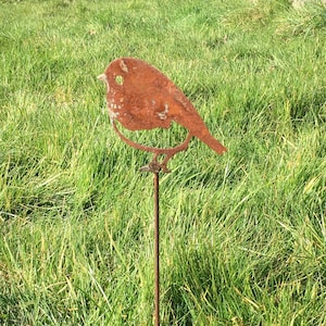 Könnte beinhalten: Ein rostiger Metall-Gartenpfahl mit einer Silhouette eines Rotkehlchens. Der Pfahl steht in einem Feld mit grünem Gras.