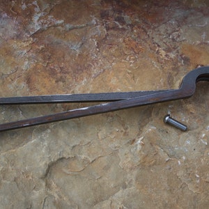 Può includere: Un paio di pinze in metallo arrugginito con un manico curvo e un piccolo perno metallico che giace accanto a loro su una superficie ruvida, marrone e testurizzata.