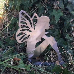 Op de afbeelding: Een zilveren metalen fee-silhouet met vleugels, zittend met gekruiste benen. De fee is geplaatst in een bed van groen gras en bladeren.