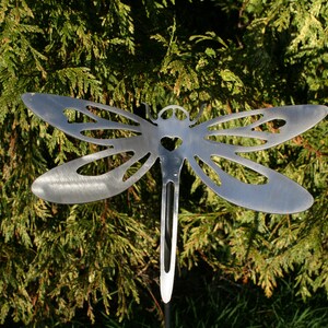 Peut inclure: Un piquet de jardin en forme de libellule en métal avec une découpe en forme de cœur au centre du corps. La libellule est argentée et a une finition brossée. Le piquet est inséré dans le sol devant un buisson vert.