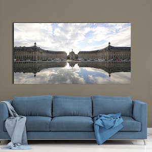 Op de afbeelding: Een groot rechthoekig canvasdoek met een Europees gebouw dat weerspiegeld wordt in water. De lucht is bewolkt met een vleugje blauw. De print hangt boven een blauwe bank met een blauwe deken. Een zwarte geometrische vloerlamp staat rechts.