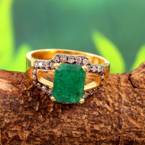 以下が含まれることがあります： 緑のエメラルドの大きな宝石が、小さなダイヤモンドで囲まれた金の指輪。指輪は茶色の木の板の上に置かれている。