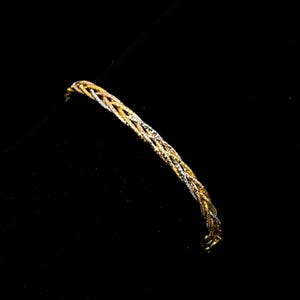 Könnte beinhalten: Ein geflochtenes Armband mit gold-, silber- und roségoldfarbenen Strängen. Das Armband besteht aus mehreren Strängen, die miteinander verwoben sind und ein strukturiertes Aussehen erzeugen. Das Armband befindet sich vor einem schwarzen Hintergrund.
