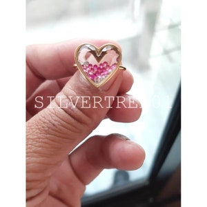 May include: A gold heart-shaped ring with a clear glass center filled with pink and clear glitter.