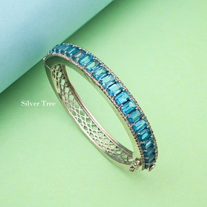 May include: A silver bangle bracelet featuring a row of light blue rectangular stones set in a silver band. The bracelet has a delicate, openwork design.