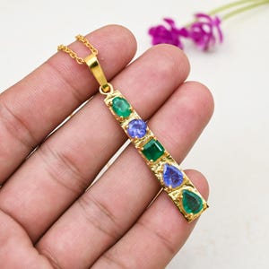 May include: A gold-colored pendant necklace featuring a vertical bar design. The bar is adorned with a series of colorful gemstones, including emeralds and tanzanites. The gemstones are in various shapes, including round, square, and teardrop. The necklace is held in a hand.