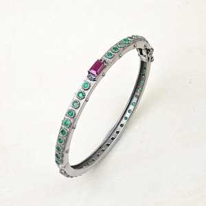 May include: A silver-coloured bangle bracelet featuring a rectangular ruby gemstone and multiple round emeralds. The hinged bracelet is decorated with small, round gemstones along its edges. The design is elegant and sophisticated, suitable for special occasions.