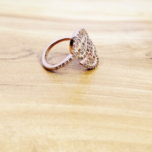 May include: A rose gold ring with a large, intricate design of sparkling clear stones. The ring is set on a light brown wooden surface.