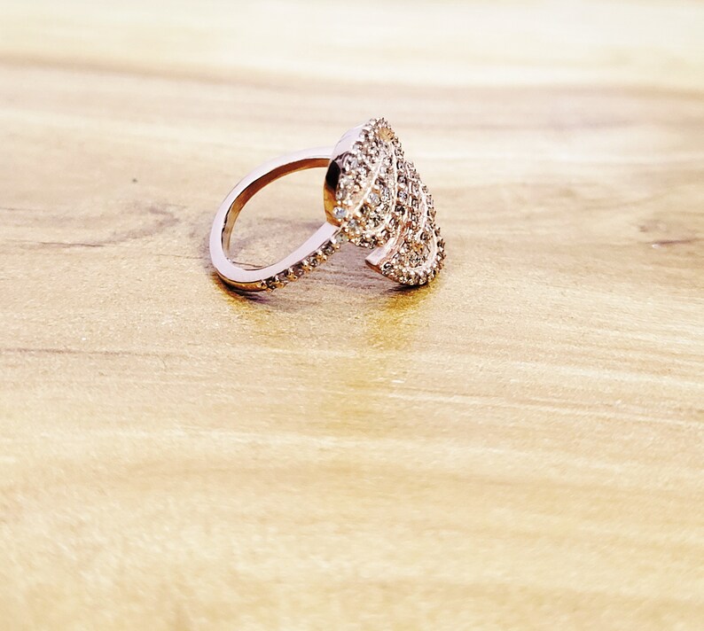 May include: A rose gold ring with a large, intricate design of sparkling clear stones. The ring is set on a light brown wooden surface.
