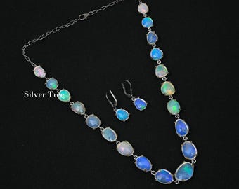 May include: A silver necklace and matching earrings set featuring oval-shaped opals in shades of blue, green, and white. The necklace and earrings are displayed on a black background. The text "Silver Tree" is visible on the left side of the image.
