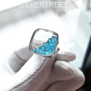 Könnte beinhalten: Ein silberner Ring mit einer quadratischen Fassung, die mit blauen Edelsteinen gefüllt ist. Der Ring liegt auf einer weißen Oberfläche.