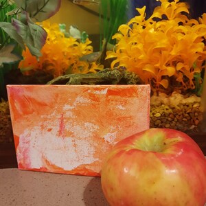 May include: A small orange and white abstract painting on canvas with a red apple in the foreground.