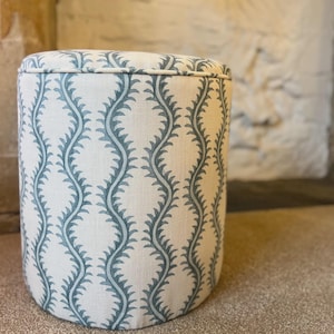 May include: A cylindrical ottoman upholstered in an off-white fabric featuring a repeating vertical pattern of blue, fern-like botanical vines. The top edge of the stool is finished with a matching fabric piping detail along the circular seam.