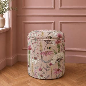 May include: A cylindrical ottoman upholstered in a light pink fabric featuring a botanical print of various wildflowers, grasses, and bees in shades of pink, white, green, and yellow. The stool has a flat, circular top and a structured, upright form.