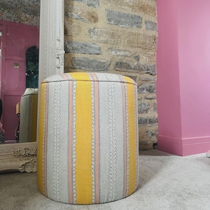 May include: A round ottoman with a striped design in yellow, pink, and white. The fabric-covered footstool has a lid. It is placed on a light-colored carpet. The background features a stone wall and a pink wall.