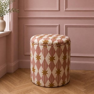 May include: A cylindrical ottoman upholstered in fabric featuring a repeating diamond pattern in muted pink and cream, accented with gold-coloured star motifs at each intersection. The stool has a flat circular top and a continuous vertical side.