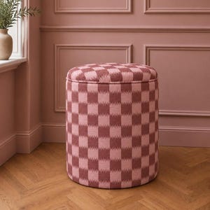 May include: A cylindrical ottoman featuring a chequered pattern in alternating shades of dusty rose and light pink. The fabric has a textured appearance with soft, blurred edges on the squares, and the top is circular with piped detailing.
