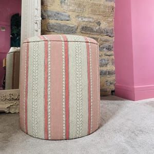May include: A cylindrical ottoman upholstered in fabric featuring vertical stripes in shades of dusty pink and cream. The pattern consists of alternating solid pink bands and cream sections decorated with a delicate, dark-dotted leaf vine motif.