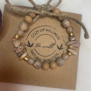 May include: A brown and pink beaded bracelet with gold accents. The bracelet is on a brown card with the text "Cozy Up Wellness Be Well Natural Skin Care".