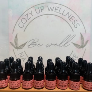 May include: A collection of small glass bottles with black droppers, filled with essential oils. The bottles are labeled with the brand name "Cozy Up Wellness" and various essential oil names, including Lavender, Rosemary, Bergamot, and Eucalyptus.