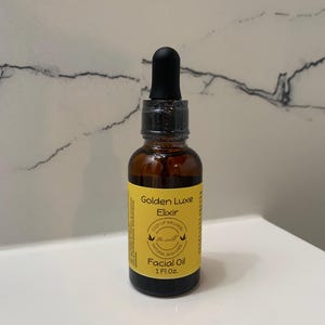 May include: A brown glass bottle of Golden Luxe Elixir facial oil with a black dropper. The bottle has a yellow label with the text "Golden Luxe Elixir" and "Facial Oil 1 Fl Oz."