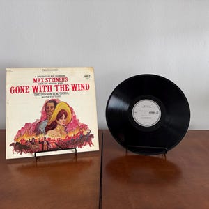 May include: A vintage vinyl record album of "Gone With The Wind" by Max Steiner. The album cover features a colorful illustration of the film's characters. A black vinyl record is displayed next to the album on a black metal stand.