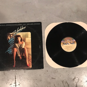May include: A vinyl record and album cover from the movie "Flashdance." The album cover is black with a photo of a woman and the movie title in a stylized font. The record is black and sits in a white sleeve.