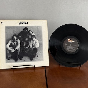 May include: A black and white photo of a band called "Rufus" on a vinyl record album cover. The band members are all wearing casual clothing and are standing in a group.