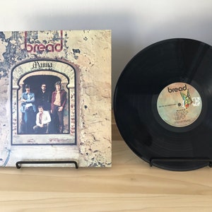 May include: A vinyl record album cover and record featuring the band Bread. The album cover is a close-up of a stone building with a window. The band members are standing in the window. The album title is "Manna" and the band's name is "Bread". The record label is black with white text and a small image of a bird.