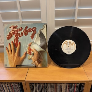 May include: Album cover for The Tubes featuring a hand with long red nails tearing away a layer of the cover. A black vinyl record is displayed next to the album cover. The album cover has the band's name in red.