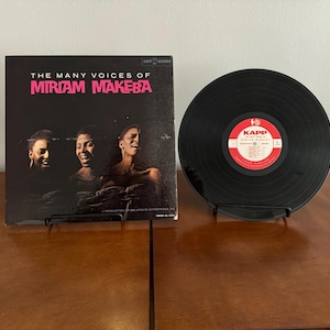 May include: Vintage vinyl record album titled "The Many Voices of Miriam Makeba" with a black record next to it. The album cover features three black and white portraits of Miriam Makeba. The record has a red and white label.
