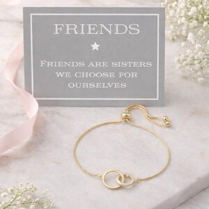 Könnte beinhalten: Ein verstellbares goldfarbenes Armband mit zwei ineinandergreifenden Kreisen, einer schlicht und einer mit kleinen klaren Steinen besetzt. Dahinter eine graue Karte mit dem weißen Text: FRIENDS, FRIENDS ARE SISTERS WE CHOOSE FOR OURSELVES.