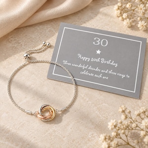 May include: A silver-tone adjustable chain bracelet featuring three interlocking rings in silver, gold, and rose gold tones. It is accompanied by a gray rectangular card with the text 30, Happy 30th Birthday, and Three wonderful decades and three rings to celebrate each one.