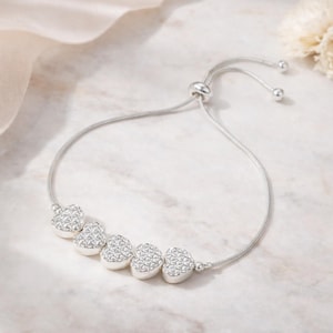 Peut inclure: Un bracelet r&eacute;glable de couleur argent&eacute;e orn&eacute; de cinq breloques en forme de c&oelig;ur avec une surface textur&eacute;e &agrave; motifs crois&eacute;s. Le bracelet est compos&eacute; d'une fine cha&icirc;ne serpent flexible et d'un m&eacute;canisme coulissant pour ajuster la taille.