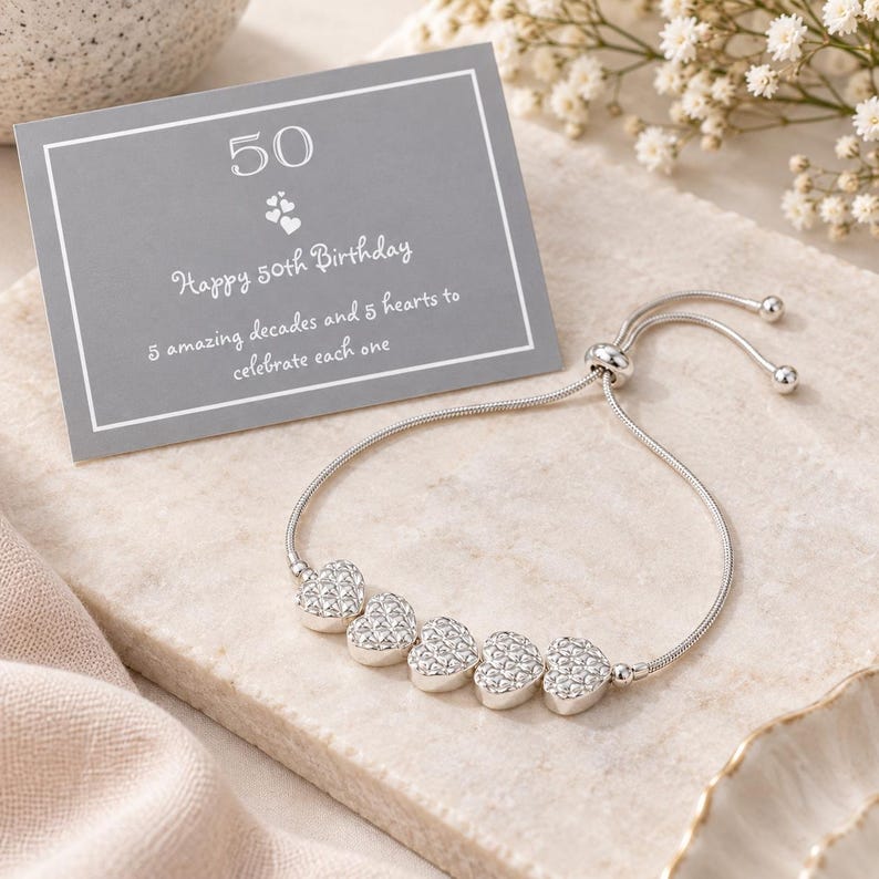 Peut inclure: Un bracelet r&eacute;glable argent&eacute; orn&eacute; de cinq breloques en forme de c&oelig;ur textur&eacute;. &Agrave; c&ocirc;t&eacute; se trouve une carte rectangulaire grise avec le texte blanc : 50, Happy 50th Birthday, 5 amazing decades and 5 hearts to celebrate each one, et une ic&ocirc;ne de c&oelig;ur.