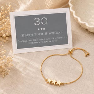 May include: A gold-toned adjustable bracelet featuring three small star charms, accompanied by a rectangular card with a gray background. The card displays the text 30, three stars, HAPPY 30TH BIRTHDAY, and 3 AMAZING DECADES AND 3 STARS TO CELEBRATE EACH ONE.