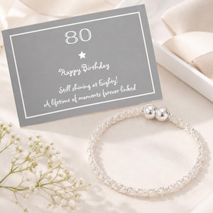 May include: A silver bracelet with two round silver beads and a gray card that reads "80 Happy Birthday Still shining at Eighty! A lifetime of moments forever linked."